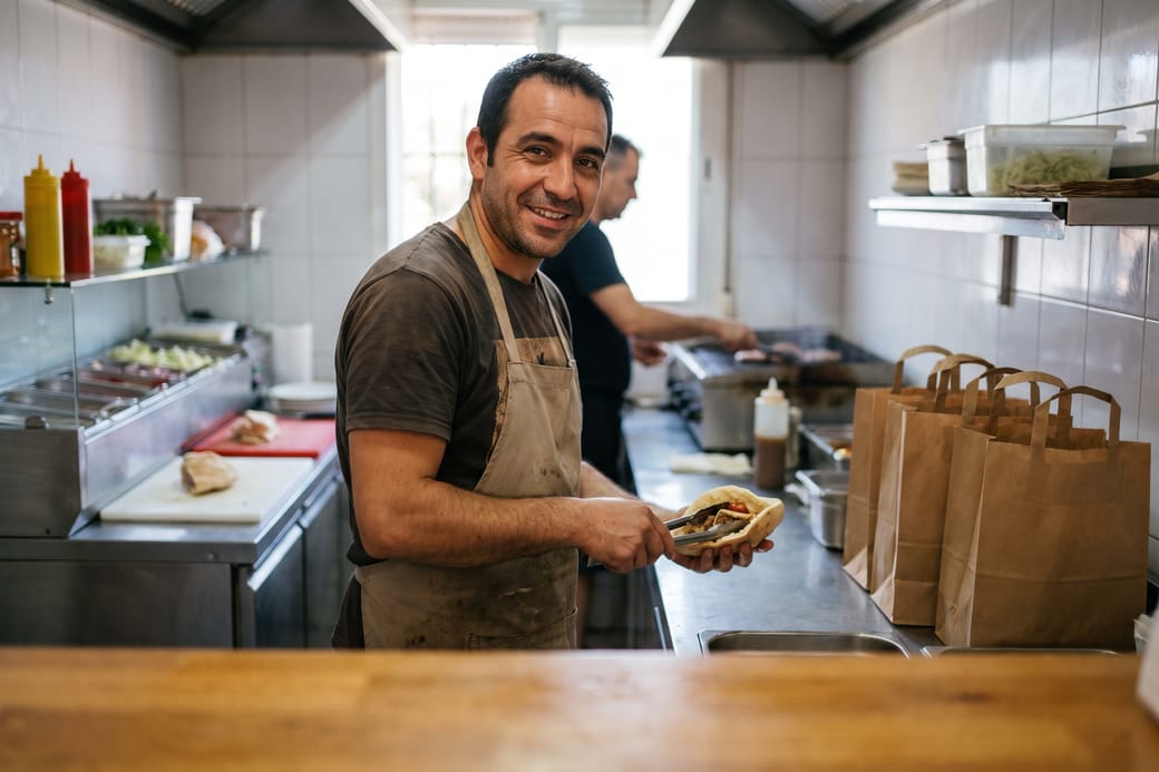 Dueño de un local de comida para llevar preparando un kebab, con las bolsas de los pedidos en el mostrador